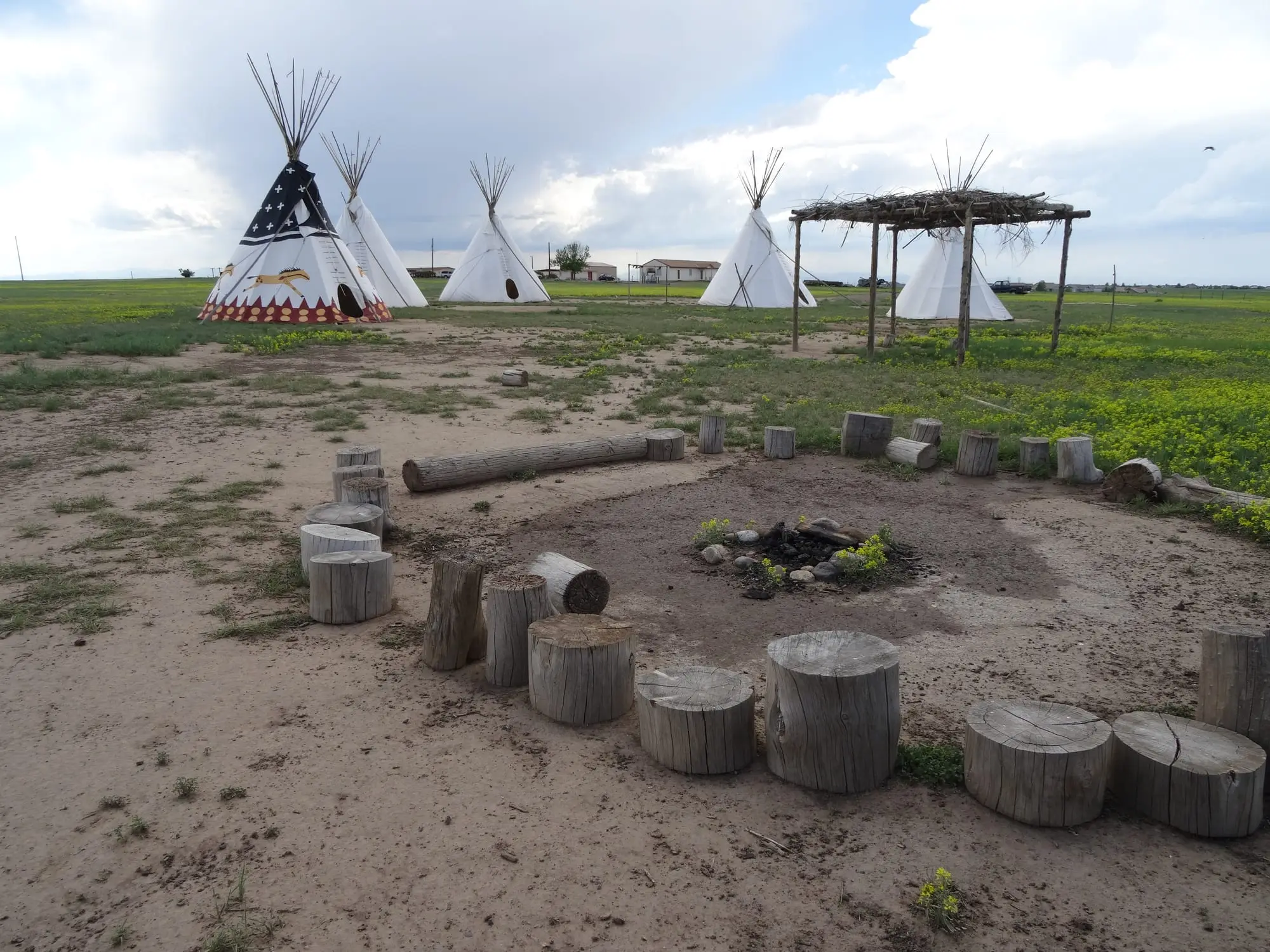 Several white tipis on open dry ground beside a ring of log stumps and a fire pit, under a blue sky with large clouds