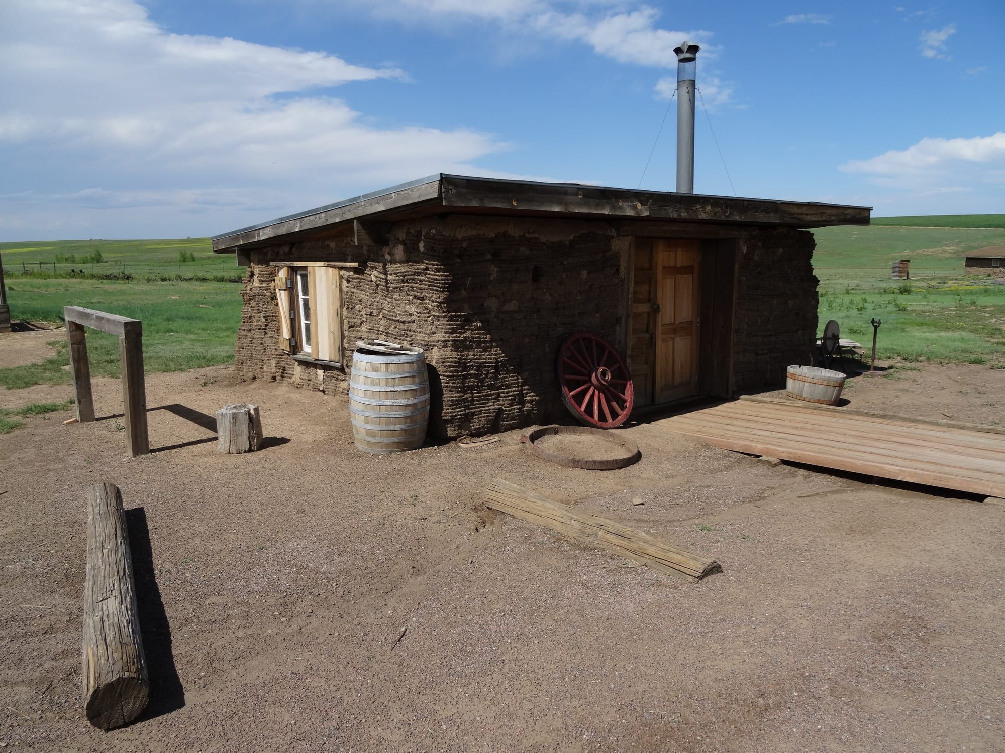 A low house of earth blocks with a flat roof and a metal stovepipe rising from it, a barrel and a wooden bench in front, on bare dry ground under a blue sky